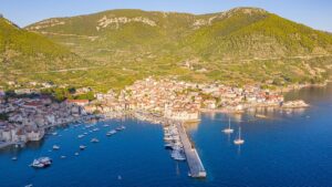 Aerial view of Komiža town and harbor on Vis Island, Croatia, with boats docked along the coast and lush green hills in the background